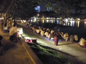 Đây là một khung cảnh vào ban đêm tại một địa điểm công cộng, có khả năng là bờ hồ ở Hà Nội. Rất đông người, chủ yếu là giới trẻ, đang tụ tập dọc theo bờ hồ. Họ ngồi trên những chiếc ghế nhựa thấp màu xanh, một hình ảnh quen thuộc của văn hóa "cà phê bệt" hay "trà chanh vỉa hè". Mọi người đang ngồi thành từng nhóm, hướng mặt ra hồ hoặc trò chuyện với nhau, tận hưởng không khí mát mẻ của buổi tối. Phía trước là mặt hồ rộng lớn, phản chiếu ánh đèn lung linh từ các tòa nhà và đèn đường ở phía xa. Dọc bờ hồ có nhiều cây xanh. Ở phía tiền cảnh, có một người đang chuẩn bị gì đó bên cạnh một chiếc hộp có đèn và vài chiếc thùng màu đỏ, có thể là một người bán hàng rong phục vụ đồ uống hoặc đồ ăn nhẹ cho đám đông. Không có văn bản hay biển hiệu nào có thể nhận dạng được trong ảnh.。（日本語訳）。これはハノイの湖畔と思われる、ある公共の場所での夜の光景です。主に若者を中心とした多くの人々が、湖畔に沿って集まっています。彼らは青い低いプラスチックの椅子に座っており、これは「路上コーヒー」や「歩道レモンティー」文化のおなじみの光景です。人々はグループになって座り、湖の方を向いたり、お互いに会話したりして、夜の涼しい空気を楽しんでいます。前方には広大な湖面が広がり、遠くの建物や街灯からのきらめく光を反射しています。湖畔に沿って多くの緑の木々があります。前景には、ライト付きの箱といくつかの赤い容器の横で何かを準備している人がいます。おそらく、人々に飲み物や軽食を提供する露天商でしょう。写真の中には、識別できる文字や看板はありません。