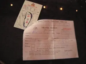 Hình ảnh chụp cận cảnh các giấy tờ du lịch đặt trên một bề mặt tối màu, có thể là mặt bàn. Đối tượng chính là một chiếc vé du lịch (TRAVEL TICKET) và một tờ giấy dán (sticker).。Trên vé có các thông tin được ghi lại như sau:。- Tên công ty: 'Hanoi Toserca - Open Tour - Sinh Cafe'.。- Tiêu đề: 'TRAVEL TICKET'.。- Mã vé: 'AB 071609' được in màu đỏ.。- Các mục như 'Itinerary' (Hành trình), 'Name' (Tên), 'Nationality' (Quốc tịch) được điền bằng tay nhưng chữ viết mờ, khó đọc. Dựa trên bối cảnh địa lý Ninh Bình, mục 'Itinerary' có khả năng cao là 'Hoa Lư - Tam Cốc'.。- Địa chỉ văn phòng: '18 Luong Van Can Str., Hanoi. OPEN TOUR OFFICE'.。- Số điện thoại: 'Tel: 8287552 - 9286631'.。- Ghi chú ở cuối vé bằng tiếng Anh đề cập đến các điều khoản về giờ khởi hành và chính sách hủy vé.。Bên cạnh vé là một tờ giấy dán có logo và tên công ty 'OPEN TOUR - SINH CAFE', trên đó có in một sơ đồ trông giống bản đồ hoặc sơ đồ ghế ngồi. Khung cảnh cho thấy đây là các vật dụng chuẩn bị cho một chuyến đi, có thể là một tour du lịch đến Ninh Bình khởi hành từ Hà Nội.。（日本語訳）。暗い色の表面、おそらくテーブルの上に置かれた旅行書類のクローズアップ写真。主な被写体は、旅行チケット（TRAVEL TICKET）とステッカーです。チケットには以下の情報が記載されています：。- 会社名：「Hanoi Toserca - Open Tour - Sinh Cafe」。- タイトル：「TRAVEL TICKET」。- チケット番号：「AB 071609」は赤色で印刷されています。- 「Itinerary」（旅程）、「Name」（名前）、「Nationality」（国籍）などの項目は手書きで記入されていますが、文字がかすれていて読みにくいです。ニンビンの地理的背景に基づくと、「Itinerary」の項目は「ホアルー - タムコック」である可能性が高いです。- オフィス住所：「18 Luong Van Can Str., Hanoi. OPEN TOUR OFFICE」。- 電話番号：「Tel: 8287552 - 9286631」。- チケットの最後にある英語の注記は、出発時間とキャンセルポリシーに関する条項に言及しています。チケットの隣には、「OPEN TOUR - SINH CAFE」という会社のロゴと名前が入ったステッカーがあり、その上には地図または座席表のような図が印刷されています。この光景は、旅行の準備品であることを示しており、おそらくハノイから出発するニンビンへの観光ツアーのものでしょう。