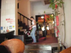 Bức ảnh được chụp bên trong một không gian thương mại, có khả năng là sảnh của một khách sạn, nhà nghỉ hoặc nhà hàng. Ở trung tâm, một người phụ nữ mặc áo ba lỗ màu trắng và quần jean xanh đang di chuyển, dường như đang bước xuống một bậc thềm. Phía bên trái, trên một bức tường ốp gạch trắng, có một biển báo màu đỏ ghi chữ "WIFI...", phần văn bản còn lại bên dưới không thể đọc rõ. Dưới dòng chữ là một số logo nhỏ, mờ. Phía sau người phụ nữ là một cầu thang với lan can tối màu. Ở bên phải của bức ảnh, một cây cảnh, có thể là cây tre hoặc trúc, được trang trí bằng nhiều dải ruy băng màu đỏ. Xa hơn ở phía sau là một lối vào dẫn đến một khu vực khác được chiếu sáng.。（日本語訳）。写真は商業施設内で撮影されたもので、ホテル、旅館、またはレストランのロビーである可能性があります。中央では、白いタンクトップと青いジーンズを着用した女性が移動しており、段差を降りているように見えます。左側には、白いタイル張りの壁に「WIFI...」と書かれた赤い看板があり、その下の残りのテキストははっきりと読み取れません。文字の下には、いくつかの小さくてぼやけたロゴがあります。女性の後ろには、暗い色の手すりが付いた階段があります。写真の右側には、観葉植物、おそらく竹か笹があり、たくさんの赤いリボンで飾られています。さらに奥には、別の照らされたエリアに通じる入り口があります。