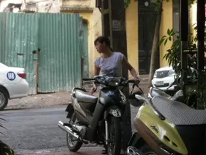 Bối cảnh là một con phố hoặc một con hẻm. Một người phụ nữ trẻ đang đứng cạnh một chiếc xe máy màu đen, tay cầm ghi đông và một chiếc mũ bảo hiểm, có vẻ như đang chuẩn bị di chuyển xe. Ở phía trước, có một chiếc xe máy tay ga màu vàng với chữ "Attila" trên thân xe. Bên trái là một chiếc ô tô màu trắng, trên thân xe có một hình tròn màu xanh dương với số "118". Phía sau người phụ nữ là một hàng rào bằng tôn lượn sóng màu xanh lá cây, có thể là rào chắn của một công trình xây dựng. Trong hậu cảnh có thể thấy một tòa nhà tường vàng và một chiếc ô tô khác đang đỗ. Trên một cây cột có một tấm biển nhỏ nhưng chữ viết không thể đọc rõ.。（日本語訳）。背景は通り、または路地です。若い女性が黒いバイクの隣に立っており、ハンドルとヘルメットを手に持ち、バイクを動かす準備をしているようです。前方には、車体に「Attila」という文字が入った黄色のスクーターがあります。左側には白い車があり、車体には「118」という数字が入った青い円が描かれています。女性の後ろには緑色の波板のフェンスがあり、建設現場の囲いである可能性があります。背景には、黄色い壁の建物と、駐車している別の車が見えます。柱には小さな看板がありますが、文字ははっきりと読み取れません。