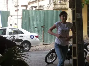 Bối cảnh là một con phố hoặc một con hẻm ở khu vực đô thị. Ở phía trước, một cô gái trẻ mặc áo phông trắng có họa tiết và quần jean xanh đang đi bộ. Phía sau cô gái là một chiếc xe máy màu đen đang đậu và một cột điện bê tông. Trên cột điện có dán nhiều tờ quảng cáo, một số nội dung có thể đọc được bao gồm: 'SỬA ... 0943.97', 'KHOAN CẮT ... 0906.174...', và các số điện thoại khác như '...2222', '0943148...'. Ở phía sau, có một chiếc ô tô sedan màu trắng, có thể là xe taxi, với số '118' được in trong một vòng tròn màu xanh ở bên hông phía sau. Có hai người đang đứng gần chiếc xe. Toàn bộ khu vực được quây lại bởi một hàng rào tôn màu xanh lá cây, có thể là một công trường xây dựng. Các tòa nhà xung quanh trông cũ kỹ. Trên tường của một tòa nhà có một biển số nhà nhỏ màu đen, có thể là số '114'.。（日本語訳）。背景は都市部のある通り、または路地です。手前には、模様の入った白いTシャツと青いジーンズを履いた若い女性が歩いています。女性の後ろには、黒いバイクが駐車しており、コンクリートの電柱があります。電柱には多くの広告チラシが貼られており、読み取れる内容には「SỬA ... 0943.97」、「KHOAN CẮT ... 0906.174...」、そして「...2222」、「0943148...」などの他の電話番号が含まれています。後方には、白いセダン車があり、おそらくタクシーで、後部側面に青い円の中に「118」という数字が印刷されています。車の近くには二人の人が立っています。エリア全体は緑色のトタンのフェンスで囲まれており、おそらく建設現場でしょう。周りの建物は古びて見えます。ある建物の壁には、黒い小さな家屋番号プレートがあり、おそらく「114」という番号です。