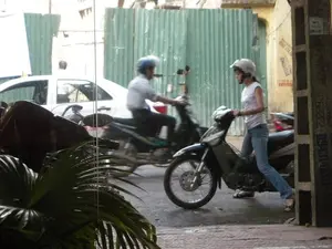 Trên cột bê tông và bức tường phía sau có dán nhiều tờ quảng cáo, một hình ảnh đặc trưng của đường phố Việt Nam. Một số nội dung có thể đọc được bao gồm 'SCA', '0943.97', và các số điện thoại khác dùng cho dịch vụ 'KHOAN CẮT BÊ TÔNG', tuy nhiên nhiều chi tiết đã bị mờ.。Bối cảnh là một con đường hoặc con hẻm trong đô thị, mặt đường ướt như vừa sau cơn mưa. Quang cảnh được chụp từ bên trong một ngôi nhà nhìn ra ngoài qua cửa kính.。Hoạt động chính diễn ra trên đường phố. Ở phía trước, một người phụ nữ mặc áo phông trắng, quần jean xanh và đội mũ bảo hiểm màu trắng đang dắt một chiếc xe máy (xe số) màu đen trên vỉa hè. Ở phía sau, một người đàn ông mặc áo sơ mi trắng, đội mũ bảo hiểm màu xanh đang lái một chiếc xe máy khác, hình ảnh bị nhòe đi do đang di chuyển. Ngoài ra, có một chiếc ô tô màu trắng đang đỗ ở phía sau.。Các vật thể đáng chú ý bao gồm nhiều xe máy (xe máy), mũ bảo hiểm (mũ bảo hiểm), một chiếc ô tô, một hàng rào bằng tôn lượn sóng màu xanh lá cây và một chậu cây cảnh ở tiền cảnh.。（日本語訳）。後ろのコンクリートの柱と壁には、ベトナムの街の典型的な光景である多くの広告が貼られています。読み取れる内容には、「SCA」、「0943.97」、および「コンクリート穿孔・切断」サービス用のその他の電話番号が含まれますが、多くの詳細はぼやけています。背景は都市の道または路地で、路面は雨上がりのように濡れています。光景は、家の中からガラス窓を通して外を眺めるように撮影されています。主な活動は通りで行われています。手前では、白いTシャツ、青いジーンズを着用し、白いヘルメットをかぶった女性が、歩道で黒いバイク（カブ型バイク）を押しています。後方では、白いシャツを着て青いヘルメットをかぶった男性が別のバイクを運転しており、移動中のためその姿はぼやけています。さらに、後方には白い車が一台駐車しています。注目すべき物体には、多くのバイク（xe máy）、ヘルメット（mũ bảo hiểm）、一台の車、緑色の波板トタンのフェンス、そして前景にある観葉植物の鉢が含まれます。