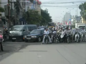Bức ảnh ghi lại cảnh giao thông đông đúc trên một con đường nhựa trong một khu đô thị ở Việt Nam. Rất nhiều người đi xe máy đang dừng lại, xếp thành hàng ngang trên đường, có lẽ đang chờ tín hiệu giao thông. Hầu hết người điều khiển xe máy đều đội mũ bảo hiểm. Xen lẫn trong dòng phương tiện còn có một chiếc ô tô con màu sẫm và một chiếc xe van màu bạc. Ở phía bên trái là các tòa nhà phố và một mạng lưới dây điện phức tạp, chằng chịt phía trên. Phía xa trong hậu cảnh, có thể nhìn thấy các tòa nhà khác, cột đèn đường và nhiều phương tiện hơn. Mặc dù có các biển hiệu trên các tòa nhà, hình ảnh bị mờ nên không thể đọc được nội dung.。（日本語訳）。この写真は、ベトナムのある都市部のアスファルト道路における混雑した交通風景を記録したものです。非常に多くのバイクに乗った人々が停止し、道路上で横一列に並んでおり、おそらく交通信号を待っています。バイクの運転手のほとんどはヘルメットを着用しています。車両の流れの中には、濃い色の乗用車1台と銀色のバン1台も混じっています。左側には商店が連なる建物と、その上には複雑に絡み合った電線の網があります。遠くの背景には、他の建物、街灯、さらに多くの車両が見えます。