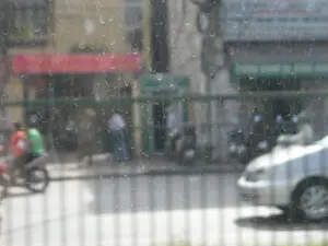 Bức ảnh được chụp từ bên trong nhìn ra ngoài qua một tấm kính mờ, có thể là cửa sổ bị bẩn hoặc dính các vệt nước khô. Khung cảnh bên ngoài là một con đường phố. Ở phía bên trái, có một chiếc xe máy đang chạy với hai người ngồi trên, một người mặc áo màu cam và người kia mặc áo màu xanh lá cây. Ở phía bên phải, có thể thấy phần đầu của một chiếc ô tô màu sáng. Phía bên kia đường là các tòa nhà, có vẻ là các cửa hàng, với các mái hiên màu đỏ và xanh lá cây. Có các biển hiệu nhưng chữ viết trên đó hoàn toàn không thể đọc được do hình ảnh bị mờ. Toàn bộ khung cảnh mô tả một hoạt động giao thông và sinh hoạt thường nhật trên một con phố.。（日本語訳）。写真は、汚れた窓か乾いた水滴の跡がついている曇りガラスを通して、内側から外を見て撮られたものです。外の景色は街路です。左側には、2人乗りのバイクが走っており、1人はオレンジ色の服を、もう1人は緑色の服を着ています。右側には、明るい色の車の前部が見えます。道の向こう側には建物があり、店のようで、赤と緑のひさしが付いています。看板がありますが、画像がぼやけているため、そこに書かれた文字は全く読めません。全景は、ある街路での交通と日常生活の様子を描写しています。