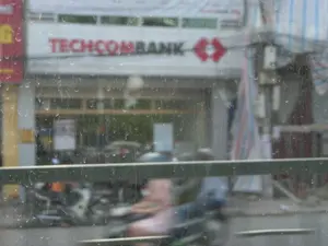 Bức ảnh được chụp qua một ô cửa kính dính đầy hạt mưa, nhìn ra một con đường ở đô thị, có thể là tại Ninh Bình. Bên kia đường là một tòa nhà có biển hiệu lớn màu đỏ với dòng chữ 'TECHCOMBANK'. Đây dường như là một chi nhánh ngân hàng. Phía dưới, trên đường phố, có thể thấy bóng người đang điều khiển xe máy di chuyển, nhưng hình ảnh bị mờ do phương tiện chụp ảnh đang chuyển động và do trời mưa. Khung cảnh chung cho thấy một hoạt động giao thông thường ngày trên đường phố Việt Nam trong một ngày mưa.。（日本語訳）。写真は、雨粒がたくさんついたガラス窓越しに撮影されたもので、都市の通りを眺めている。場所はニンビンかもしれない。通りの向こう側には、「TECHCOMBANK」という文字が書かれた大きな赤い看板のある建物がある。これは銀行の支店のようだ。全体的な光景は、雨の日のベトナムの路上における日常的な交通の様子を示している。