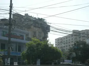 Đây là một khung cảnh đường phố tại một đô thị ở Việt Nam. Bức ảnh cho thấy sự pha trộn giữa các công trình kiến trúc cũ và mới. Ở hậu cảnh là một tòa nhà tập thể cũ, tường đã xuống cấp, và bên cạnh là một tòa nhà chung cư hiện đại hơn. Phía trước là một tòa nhà hai tầng có vẻ là một văn phòng hoặc cơ quan. Một đặc điểm nổi bật là mạng lưới dây điện và cáp viễn thông chằng chịt trên bầu trời, cùng với một cột điện lớn ở bên trái. Cây xanh tươi tốt mọc ở tiền cảnh. Trên tòa nhà hai tầng có treo một băng rôn màu đỏ với chữ vàng, nội dung được ghi lại như sau: 'NHIỆT LIỆT CHÀO MỪNG KỶ NIỆM 63 NĂM CÁCH MẠNG THÁNG 8 VÀ QUỐC KHÁNH 2/9'. Bên dưới có một biển hiệu màu xanh với các từ có thể đọc được như 'CÔNG TY...', 'THANH TOÁN CƯỚC PHÍ', 'GIẢI QUYẾT KHIẾU NẠI'. Trên nóc tòa nhà cũ có các bồn nước inox và ăng-ten. Khung cảnh không cho thấy hoạt động cụ thể của con người mà chủ yếu là cảnh quan đô thị.。（日本語訳）。これはベトナムのある都市の街角の風景です。この写真は、古い建築物と新しい建築物が混在している様子を示しています。背景には、壁が劣化した古い集合住宅があり、その隣にはより近代的なマンションがあります。手前には、オフィスか役所と思われる2階建ての建物があります。目立つ特徴は、空に張り巡らされた電線や通信ケーブルの網と、左側にある大きな電柱です。前景には青々とした木々が生い茂っています。2階建ての建物には、黄色い文字の入った赤い横断幕が掲げられており、その内容は次のように記されています。「8月革命及び9月2日建国記念日63周年を熱烈に歓迎します」。その下には、「会社...」、「料金支払い」、「苦情解決」といった読み取れる言葉が書かれた青い看板があります。古い建物の屋上には、ステンレス製の給水タンクとアンテナがあります。この風景は、特定の人の活動を示しているのではなく、主に都市景観です。