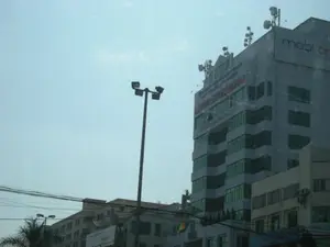 Bức ảnh chụp một khung cảnh đường phố ở khu vực đô thị. Nổi bật là một tòa nhà văn phòng cao tầng, hiện đại. Trên đỉnh tòa nhà có một biển hiệu lớn ghi chữ 'mobifone'. Bên dưới biển hiệu này có các dòng chữ khác, trong đó có thể nhận ra dòng chữ 'CÔNG TY THÔNG TIN DI ĐỘNG' và một dòng chữ màu đỏ khó đọc hơn. Tòa nhà có nhiều cửa sổ kính màu xanh lá. Trên nóc nhà được lắp đặt rất nhiều ăng-ten và các thiết bị thu phát sóng viễn thông. Ở phía trước, có một cột đèn đường cao. Phía dưới là các tòa nhà khác và hệ thống dây điện chằng chịt. Bầu trời trong xanh. Khung cảnh cho thấy đây là một khu vực thương mại hoặc trung tâm thành phố, với sự hiện diện của một công ty viễn thông lớn.。（日本語訳）。写真は都市部の街の風景を撮影したものです。目立っているのは、モダンな高層オフィスビルです。ビルの最上部には「mobifone」と書かれた大きな看板があります。この看板の下には他の文字列があり、その中には「CÔNG TY THÔNG TIN DI ĐỘNG」という文字列と、それより読みにくい赤色の文字列が認識できます。ビルには緑色のガラス窓がたくさんあります。屋上には、たくさんのアンテナや通信送受信装置が設置されています。手前には、高い街灯が一本あります。その下には他のビルや、張り巡らされた電線があります。空は青く澄んでいます。この風景は、大手通信会社の存在から、ここが商業地域または都心部であることを示しています。