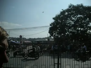 Bức ảnh chụp một cảnh đường phố đông đúc ở Việt Nam, có thể là tại Ninh Bình, được nhìn qua cửa sổ của một phương tiện. Khung cảnh cho thấy tình trạng giao thông tấp nập với rất nhiều xe máy đang di chuyển hoặc dừng lại trên đường. Nhiều người điều khiển xe máy, một số có đội mũ bảo hiểm. Ở hậu cảnh, có thể nhìn thấy ít nhất hai chiếc xe buýt và các tòa nhà. Bầu trời trong xanh và có nhiều dây điện chằng chịt vắt ngang. Phía bên phải có một cây lớn, rậm rạp. Ở tiền cảnh, có một hàng rào sắt và một phần đầu của một người ở góc trái, cho thấy góc chụp từ bên trong xe. Có các biển báo trên đường nhưng chữ viết không thể nhận dạng được.。（日本語訳）。この写真は、ベトナムの、おそらくはニンビンでの混雑した街の風景を、ある乗り物の窓越しに見たものです。その風景は、非常に多くのバイクが道路上を移動または停止している、交通が混雑した状況を示しています。多くの人がバイクを運転しており、一部はヘルメットを着用しています。背景には、少なくとも2台のバスと複数の建物が見えます。空は青く澄んでおり、多くの電線が複雑に交差しています。右側には、大きくて生い茂った木があります。前景には、鉄の柵と左隅に人の頭の一部があり、車内からの撮影アングルであることを示しています。道路には標識がありますが、文字は識別できません。