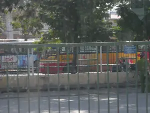 Bản ghi chép văn bản: Có một biển báo màu xanh với chữ trắng ghi: "KHU VỰC CẤM XE ÔM - HÀNG RONG HOẠT ĐỘNG". Trên thân một chiếc xe buýt, có thể đọc được một phần chữ "DAEWOO".。Mô tả cảnh vật và hoạt động: Bức ảnh chụp một khung cảnh đường phố, có thể là gần một bến xe hoặc khu vực công cộng. Ở phía trước là một hàng rào sắt màu xanh lá cây. Phía sau hàng rào, một người công an mặc sắc phục màu xanh lá, đội mũ bảo hiểm, đang đứng làm nhiệm vụ cạnh một chiếc xe đạp. Gần đó là biển báo cấm xe ôm và hàng rong hoạt động. Một chiếc xe buýt lớn có màu đỏ, vàng và trắng đang đỗ hoặc di chuyển chậm. Xung quanh có nhiều cây xanh và một phần của một tòa nhà ở phía sau. Bức ảnh dường như được chụp từ bên trong một phương tiện đang di chuyển, nhìn qua cửa sổ.。（日本語訳）。テキストの記録：白い文字で「バイクタクシー・露天商 活動禁止区域」と書かれた青い看板があります。バスの車体には、「DAEWOO」という文字の一部が読み取れます。手前には緑色の鉄柵があります。柵の後ろでは、緑色の制服を着てヘルメットをかぶった警察官が、自転車の隣で任務に就いています。その近くに、バイクタクシーと露天商の活動を禁止する看板があります。赤、黄、白の大きなバスが停車しているか、ゆっくりと移動しています。周囲には多くの緑の木々があり、後方には建物の一部が見えます。写真は移動中の乗り物の内側から、窓越しに撮影されたようです。