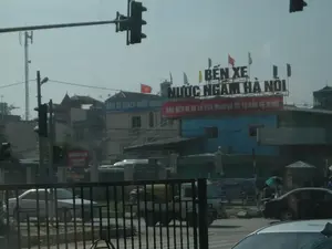 Đây là khung cảnh đường phố bên ngoài một bến xe ở Việt Nam. Có nhiều biển hiệu có thể đọc được.。Chữ viết trên các biển hiệu:。- Trên nóc một tòa nhà có biển hiệu lớn với các chữ cái màu trắng: "BẾN XE NƯỚC NGẦM".。- Ngay bên dưới là một biểu ngữ màu đỏ với chữ trắng: "VÀO BẾN ĐI XE LÀ VĂN MINH VÀ ĐỂ TỰ BẢO VỆ MÌNH".。- Trên một tòa nhà khác ở bên trái có biển hiệu: "BẾN XE KHÁCH NƯỚC NGẦM".。- Phía dưới tòa nhà đó có một biển hiệu nhỏ hơn màu đỏ ghi: "KHÁCH SẠN VĂN XUÂN".。Miêu tả cảnh quan và hoạt động:。- Loại địa điểm: Đây là khu vực bên ngoài một bến xe khách lớn, có nhiều tòa nhà, đường sá và phương tiện qua lại.。- Hoạt động: Giao thông đang diễn ra trên đường với ô tô và một chiếc xe tải nhỏ. Một người đi bộ đang che ô màu xanh lá cây. Không khí chung cho thấy sự nhộn nhịp của một khu vực cửa ngõ giao thông.。- Các vật thể trong ảnh: Nhiều tòa nhà san sát, các biển hiệu quảng cáo, cột đèn giao thông, một cột ăng-ten viễn thông cao, nhiều lá cờ trang trí nhiều màu và một lá cờ Việt Nam (cờ đỏ sao vàng). Ngoài ra còn có ô tô, xe tải và hàng rào sắt ở tiền cảnh.。（日本語訳）。これはベトナムのあるバスターミナルの外の街の風景です。読み取れる看板が多数あります。看板の文字：。- ある建物の屋上には、白い文字で「BẾN XE NƯỚC NGẦM」と書かれた大きな看板があります。- そのすぐ下には、白い文字で「VÀO BẾN ĐI XE LÀ VĂN MINH VÀ ĐỂ TỰ BẢO VỆ MÌNH」（ターミナルに入って乗車することは文明的であり、自分自身を守るためです）と書かれた赤い横断幕があります。- 左側の別の建物には、「BẾN XE KHÁCH NƯỚC NGẦM」という看板があります。- その建物の下には、「KHÁCH SẠN VĂN XUÂN」と書かれた小さな赤い看板があります。風景と活動の描写：。- 場所の種類：ここは大きな旅客バスターミナルの外のエリアで、多くの建物、道路、行き交う乗り物があります。- 活動：道路では乗用車と小型トラックが通行しています。歩行者が一人、緑色の傘をさしています。全体の雰囲気は、交通の玄関口としての賑わいを示しています。- 写真の中の物体：密集した多くの建物、広告看板、信号機の柱、高い通信アンテナの柱、多くの色とりどりの装飾旗、そしてベトナム国旗（金星紅旗）が一つあります。その他、前景には乗用車、トラック、鉄の柵があります。