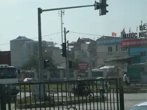 Đây là một khung cảnh đường phố tại một khu vực đô thị ở Việt Nam, có thể là gần một bến xe. Trong ảnh có các biển hiệu với nội dung: "BẾN XE KHÁCH NƯỚC NGẦM", "VÀO BẾN ĐI XE LÀ VĂN MINH", "KHÁCH SẠN VẠN XUÂN", và "KHU VỆ SINH WC". Ngoài ra còn có một biển hiệu lớn khác ghi "BẾN... NƯỚC NG...".。Cảnh vật cho thấy một ngã tư với cột đèn tín hiệu giao thông ở trung tâm. Xung quanh là các tòa nhà cao tầng, và phía xa có một cột tháp viễn thông. Cờ Việt Nam được treo trên nóc một tòa nhà. Hoạt động chính là giao thông và sinh hoạt thường ngày. Có nhiều phương tiện như xe tải, xe buýt và xe máy. Một số người đang đi bộ, trong đó có hai người phụ nữ đang che ô (một màu hồng, một màu xanh lá) để tránh nắng. Một vài người khác đang ngồi trên xe máy. Phía trước có một hàng rào sắt.。（日本語訳）。これはベトナムの都市部における街の風景で、おそらくバスターミナルの近くでしょう。写真には、「NƯỚC NGẦM（ヌックガム）旅客バスターミナル」、「ターミナルに入って乗車することは文明的な行為です」、「VẠN XUÂN（ヴァン・スアン）ホテル」、そして「WCトイレエリア」という内容の看板があります。その他にも、「BẾN... NƯỚC NG...」と書かれた大きな看板がもう一つあります。風景は、中央に信号機の柱がある交差点を示しています。周りには高層ビルがあり、遠くには通信塔があります。ベトナム国旗が建物の屋上に掲げられています。主な活動は交通と日常生活です。トラック、バス、バイクなど多くの乗り物があります。何人かの人が歩いており、その中には日差しを避けるために傘（一つはピンク、もう一つは緑）をさしている二人の女性がいます。他の数人はバイクに乗っています。手前には鉄の柵があります。