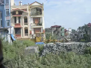 Bức ảnh chụp một khu vực dân cư ở Ninh Bình, có vẻ như đang trong quá trình xây dựng hoặc phát triển. Ở phía trước, cây cối và cỏ dại mọc um tùm. Giữa khu vực cây cối này có một đống cọc bê tông cốt thép được xếp chồng lên nhau và một chiếc máy công trình màu vàng, có thể là máy xúc hoặc máy ủi. Phía sau là các ngôi nhà ống nhiều tầng với kiến trúc hiện đại, một số có ban công trồng cây cảnh và bồn nước inox trên mái. Bên trái có một lán trại tạm bợ được dựng bằng bạt. Không có hoạt động của con người được ghi lại, nhưng sự hiện diện của vật liệu và máy móc xây dựng cho thấy đây là một công trường. Trên thân máy công trình màu vàng có chữ viết nhưng không thể nhận dạng rõ.。（日本語訳）。ニンビン省のある住宅地を写した写真で、建設中あるいは開発中であるように見えます。手前には、木々や雑草が生い茂っています。この木々が生い茂るエリアの中には、積み重ねられた鉄筋コンクリート杭の山と、ショベルカーかブルドーザーと思われる一台の黄色い建設機械があります。後ろには、現代的な建築様式の多層階のチューブハウスが複数あり、いくつかは観葉植物が植えられたバルコニーや、屋根の上にステンレス製の貯水タンクを備えています。左側には、シートで建てられた仮設の小屋があります。人の活動は記録されていませんが、建設資材や建設機械があることから、ここが工事現場であることが示されています。黄色い建設機械の車体には文字が書かれていますが、はっきりと識別することはできません。