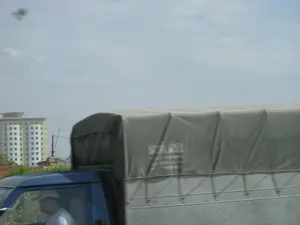 Bức ảnh chụp cảnh đường phố từ một phương tiện đang di chuyển. Ở cận cảnh là một chiếc xe tải màu xanh lam, với thùng xe được phủ bạt màu xám. Có thể thấy một người đàn ông đội mũ đang ngồi ở ghế lái. Trên tấm bạt che thùng xe có một dòng chữ màu trắng nhưng đã bị nhòe do chuyển động nên không thể đọc rõ. Ở phía sau, có một tòa nhà cao tầng màu vàng nhạt với nhiều cửa sổ. Bầu trời có mây nhẹ. Khung cảnh cho thấy hoạt động giao thông trên một con đường ở một khu vực có các công trình xây dựng.。（日本語訳）。移動中の乗り物から撮影された街の風景写真です。手前には青いトラックがあり、荷台は灰色のシートで覆われています。運転席には帽子をかぶった男性が座っているのが見えます。後方には、多くの窓がある薄黄色の高層ビルがあります。空は薄く曇っています。この光景は、建設物があるエリアの道路上の交通状況を示しています。