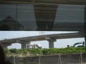 Bức ảnh ghi lại một công trường xây dựng, cụ thể là một cây cầu cạn hoặc đường cao tốc trên cao đang trong quá trình thi công. Các trụ bê tông lớn đã được dựng lên để đỡ mặt cầu. Phần mặt cầu bằng bê tông vẫn còn dang dở, với các thanh cốt thép nhô lên. Bên dưới cầu là khu vực đất trống với cây cỏ mọc um tùm. Có thể thấy các máy móc xây dựng hạng nặng, bao gồm ít nhất hai chiếc máy xúc, đang hoạt động tại công trường. Phía trước có một hàng rào lưới thép, và ở phía xa có một tòa nhà. Phần trên của bức ảnh bị tối do phản chiếu trên cửa sổ của phương tiện mà người chụp đang ở bên trong. Không có văn bản hay ký hiệu nào có thể đọc được.。（日本語訳）。この写真は建設現場を記録したもので、具体的には高架橋または高架高速道路が建設中です。大きなコンクリートの橋脚が橋桁を支えるために建てられています。コンクリートの橋桁部分はまだ未完成で、鉄筋が突き出ています。橋の下は草木が生い茂る空き地です。少なくとも2台のショベルカーを含む重建設機械が現場で稼働しているのが見えます。手前には金網のフェンスがあり、遠くには建物があります。写真の上部は、撮影者が乗っている乗り物の窓への反射により暗くなっています。読み取れる文字や記号はありません。