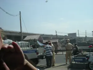 Hình ảnh ghi lại một cảnh tượng ven đường, có thể là gần một công trường xây dựng. Ở phía sau là một cây cầu vượt lớn bằng bê tông đang được thi công. Dưới mặt đất, có một vài lán trại tạm bợ. Ở tiền cảnh, một nhóm người đang đứng bên lề đường. Trong đó có hai người mặc đồng phục màu vàng của cảnh sát giao thông, đội mũ bảo hiểm. Họ đang đứng cạnh một người đàn ông mặc áo phông kẻ sọc và một chiếc xe bán tải màu trắng có vạch xanh dương. Trên thân chiếc xe bán tải có thể đọc được chữ "THÔNG". Gần đó, có một người đang ngồi trên xe máy. Bối cảnh chung cho thấy đây có thể là một chốt kiểm tra giao thông hoặc một vụ việc đang được xử lý ven đường.。（日本語訳）。画像は道路脇の光景を捉えており、おそらく建設現場の近くです。後方には、建設中の大きなコンクリート製の高架橋があります。地上には、いくつかの仮設の小屋があります。前景には、道路脇に立っている人々のグループがいます。その中には、交通警察の黄色い制服を着てヘルメットをかぶった2人の人物がいます。彼らは、縞模様のTシャツを着た男性と、青いストライプの入った白いピックアップトラックの隣に立っています。ピックアップトラックの車体には「THÔNG」という文字が読み取れます。近くには、バイクに座っている人がいます。全体的な状況から、これは交通検問所か、あるいは道路脇で処理されている事件である可能性が示唆されます。