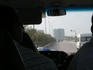 Bức ảnh được chụp từ góc nhìn của một người ngồi ở hàng ghế sau trong một chiếc ô tô đang di chuyển. Qua kính chắn gió phía trước, có thể thấy một con đường lớn, có thể là đường cao tốc hoặc quốc lộ, với nhiều phương tiện đang lưu thông. Một chiếc xe van màu trắng đang chạy ở làn đường bên phải. Dọc bên đường là hàng rào cây xanh và các cột đèn chiếu sáng. Ở phía xa, thấp thoáng bóng dáng của các tòa nhà cao tầng trong một bầu không khí có vẻ mờ ảo. Bên trong xe, có thể thấy bóng lưng của hai người ngồi phía trước; một người mặc áo sơ mi kẻ ca-rô và người còn lại mặc áo sơ mi trắng. Gương chiếu hậu phản chiếu khuôn mặt của một người đang đeo kính râm. Không có biển báo hay chữ viết nào có thể đọc được trong ảnh.。（日本語訳）。この写真は、移動中の車の後部座席に座っている人の視点から撮影されたものです。フロントガラス越しに、高速道路か国道と思われる大きな道路が見え、多くの車両が通行しています。右側の車線を白いバンが走行しています。道路沿いには緑の植え込みと街灯が並んでいます。遠くには、やや霞んだような空気の中に高層ビルの影がかすかに見えます。車内には、前に座っている2人の後ろ姿が見えます。1人はチェック柄のシャツを、もう1人は白いシャツを着ています。バックミラーにはサングラスをかけた人の顔が映っています。写真の中には、読み取れる標識や文字はありません。
