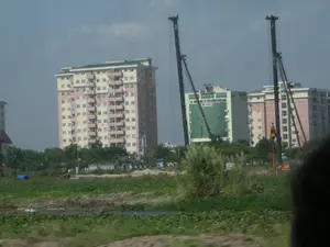 Bức ảnh chụp một khu vực ngoại ô đang trong quá trình đô thị hóa, có sự tương phản giữa tiền cảnh và hậu cảnh. Ở hậu cảnh là một công trường xây dựng với nhiều tòa nhà cao tầng. Hoạt động chính là xây dựng, được thể hiện rõ qua sự hiện diện của hai máy đóng cọc lớn màu đen. Các tòa nhà bao gồm chung cư cao tầng màu be và hồng, và một tòa nhà văn phòng hiện đại với mặt tiền bằng kính màu xanh lá cây. Trên tòa nhà văn phòng này có các dòng chữ: logo chữ "V" màu đỏ, "TỔNG CÔNG TY CÔNG NGHIỆP TÀU THỦY VIỆT NAM", và bên dưới là "CÔNG TY VẬN TẢI VIỄN DƯƠNG VINASHIN" cùng với logo "NAM TRIEU". Tiền cảnh là một khu đất trống với thảm thực vật xanh tươi, có thể là một mảnh ruộng hoặc vùng đất hoang gần công trường.。（日本語訳）。写真は都市化が進む郊外の地域を写しており、前景と後景の間にはコントラストがあります。後景には、多くの高層ビルが建ち並ぶ建設現場があります。主な活動は建設であり、それは2台の大きな黒い杭打ち機の存在によって明確に示されています。建物には、ベージュ色とピンク色の高層マンション、そして緑色のガラス張りのファサードを持つ近代的なオフィスビルが含まれています。このオフィスビルには、赤い「V」の文字のロゴ、「TỔNG CÔNG TY CÔNG NGHIỆP TÀU THỦY VIỆT NAM」、そしてその下には「NAM TRIEU」のロゴと共に「CÔNG TY VẬN TẢI VIỄN DƯƠNG VINASHIN」という文字列があります。前景は緑豊かな植生に覆われた空き地で、建設現場近くの畑か荒れ地である可能性があります。