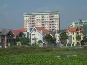 Trên tòa nhà văn phòng màu xanh lá cây ở phía sau có một biển hiệu lớn. Dòng chữ trên cùng có thể đọc được là 'CÔNG TY TNHH TƯ VẤN TÀI VIỄN DƯƠNG...', phần còn lại của tên không rõ. Bên dưới có logo và tên của ngân hàng 'Sacombank'.。Bức ảnh chụp một khu vực đô thị đang phát triển, có sự kết hợp giữa các công trình xây dựng và cảnh quan tự nhiên. Ở phía sau là các tòa nhà cao tầng, nổi bật là một tòa chung cư lớn sơn màu hồng và vàng với nhiều ban công, và tòa nhà văn phòng màu xanh lá cây đã đề cập. Ở dãy nhà gần hơn là các ngôi nhà kiểu biệt thự và nhà liền kề có mái ngói màu đỏ và xám. Một số ngôi nhà có kiến trúc hiện đại. Có một ngôi nhà đang xây dựng dở dang với phần gạch thô lộ ra ngoài. Cây xanh được trồng xung quanh các ngôi nhà. Ở tiền cảnh là một mặt nước như ao hoặc hồ, bị bao phủ gần như hoàn toàn bởi một thảm bèo tây (lục bình) màu xanh.。（日本語訳）。奥にある緑色のオフィスビルの上には、大きな看板があります。一番上の文字列は「CÔNG TY TNHH TƯ VẤN TÀI VIỄN DƯƠNG...」と読めますが、名前の残りの部分は不明です。その下には「Sacombank」銀行のロゴと名前があります。この写真は、建設物と自然景観が混在する、発展中の都市部を撮影したものです。背景には高層ビル群があり、特に目立つのは多くのバルコニーを持つピンクと黄色に塗られた大きな集合住宅と、前述の緑色のオフィスビルです。より手前の家並みには、赤色と灰色の瓦屋根を持つヴィラ風の家やタウンハウスがあります。いくつかの家は現代的な建築様式です。未加工のレンガ部分がむき出しになった、建設途中の家が一軒あります。家々の周りには緑の木々が植えられています。前景には池か湖のような水面があり、緑色のホテイアオイ（lục bình）の絨毯でほぼ完全に覆われています。