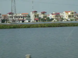 Bức ảnh ghi lại một khung cảnh ven một vùng nước rộng lớn, có thể là sông, hồ hoặc ao nuôi trồng thủy sản, tại một khu vực dân cư. Phía trước là mặt nước phẳng lặng. Ngăn cách khu dân cư và mặt nước là một bờ đất được phủ bởi thảm thực vật xanh. Ở phía sau là một dãy nhà ở với nhiều kiểu kiến trúc khác nhau. Các ngôi nhà này chủ yếu là nhà cao tầng (2-3 tầng), có ban công, mái lợp ngói đỏ, và được sơn màu trắng hoặc vàng. Nổi bật ở phía bên trái là một cột điện cao thế bằng sắt thép, và ở phía xa là một cột tháp viễn thông. Không có người hay hoạt động cụ thể nào đang diễn ra. Không có bất kỳ văn bản hay biển báo nào có thể đọc được trong ảnh.。（日本語訳）。写真は、ある住宅地にある、川、湖、あるいは養殖池かもしれない広大な水域のほとりの光景を記録しています。手前には穏やかな水面があります。住宅地と水面を隔てているのは、緑の植生で覆われた土手です。後方には、様々な建築様式の住宅が立ち並んでいます。これらの家は主に高層住宅（2〜3階建て）で、バルコニーがあり、屋根は赤い瓦で葺かれ、白または黄色に塗装されています。左側で目立っているのは鉄鋼製の高圧電柱で、遠方には通信塔があります。特定の人物や活動は行われていません。写真の中には、読み取れる文字や看板は一切ありません。