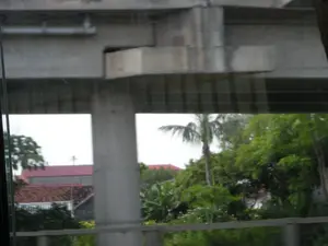 Bức ảnh được chụp từ bên trong một phương tiện đang di chuyển, nhìn ra ngoài qua cửa sổ. Khung cảnh bên ngoài bị che khuất một phần bởi một công trình cầu bê tông hoặc đường trên cao lớn với các cột trụ vững chãi. Xuyên qua các khoảng trống của cây cầu, có thể thấy một khu vực dân cư với những ngôi nhà mái ngói, một trong số đó có mái màu đỏ. Phía sau những ngôi nhà là cây cối xanh tươi, rậm rạp, nổi bật có một cây cọ (hoặc cây dừa). Một công trình ở phía xa có mái đỏ và một chi tiết trên đỉnh trông giống như cây thánh giá, gợi ý rằng đó có thể là một nhà thờ. Bầu trời có màu trắng đục, cho thấy thời tiết nhiều mây. Không có hoạt động cụ thể nào của con người được thể hiện rõ ràng.。（日本語訳）。写真は移動中の乗り物の内側から、窓を通して外を見て撮影されたものです。外の景色は、頑丈な柱を持つ大きなコンクリートの橋または高架道路の構造物によって部分的に遮られています。橋の隙間からは、瓦屋根の家々が並ぶ住宅地が見え、そのうちの一軒は赤い屋根をしています。家々の後ろには青々とした木々が茂っており、ヤシの木（またはココヤシの木）が目立っています。空は乳白色で、曇り空であることを示しています。特定の人間の活動ははっきりと見えません。