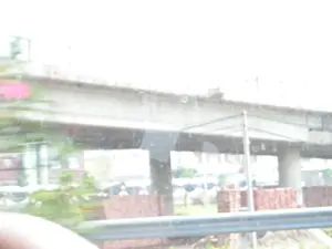 Bức ảnh bị mờ và lóa sáng, có vẻ như được chụp từ bên trong một phương tiện đang di chuyển khi nhìn qua cửa sổ. Hình ảnh cho thấy một công trình xây dựng lớn, có khả năng là một cây cầu vượt hoặc một tuyến đường bộ trên cao đang trong quá trình thi công. Cấu trúc chính bao gồm các cột trụ và dầm bê tông lớn. Bên dưới gầm cầu, có nhiều chồng gạch đỏ được xếp ngay ngắn, đặc trưng của một công trường xây dựng. Ở phía trước, có một lan can kim loại ven đường. Phông nền phía sau khá mờ, chỉ có thể nhận ra lờ mờ hình bóng của cây cối và một vài tòa nhà. Không có bất kỳ chữ viết hay biển báo nào có thể đọc được trong ảnh.。（日本語訳）。写真はぼやけて露出オーバーになっており、移動中の乗り物の車内から窓越しに撮影されたようです。画像には、大規模な建設工事が写っており、おそらく高架橋または高架道路が建設中であると思われます。主な構造物は、大きなコンクリートの柱と梁で構成されています。橋桁の下には、建設現場に特徴的な、きれいに積み上げられた赤いレンガの山が多数あります。手前には、道路脇の金属製ガードレールがあります。背景はかなりぼやけており、木々やいくつかの建物の影がかすかに認識できるだけです。写真の中には、読み取れる文字や標識は一切ありません。