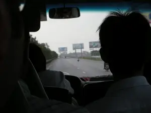 Bức ảnh được chụp từ góc nhìn bên trong một chiếc ô tô đang di chuyển trên một con đường quốc lộ hoặc đường cao tốc. Nhìn qua kính chắn gió, con đường trải dài về phía trước với một vài chiếc xe máy đang lưu thông. Dọc hai bên đường có nhiều biển quảng cáo lớn được dựng lên, tuy nhiên do khoảng cách xa và chất lượng hình ảnh, nội dung chữ viết trên các biển quảng cáo này không thể đọc được. Bên trong xe, có thể thấy rõ bóng của những người ngồi ở hàng ghế trước và sau. Ở phía trước bên phải là bóng của một người đàn ông mặc áo sơ mi trắng. Gương chiếu hậu phản chiếu hình ảnh một người đang đeo kính râm, có thể là tài xế. Khung cảnh chung cho thấy một chuyến đi trên đường bộ trong điều kiện thời tiết có vẻ u ám hoặc nhiều sương mù.。（日本語訳）。写真は、国道または高速道路を走行中の車内からの視点で撮影されたものです。フロントガラス越しに見ると、道は前方に伸びており、数台のバイクが走行しています。道路の両脇には多くの大きな広告看板が設置されていますが、距離が遠く画質も悪いため、これらの広告看板に書かれている文字の内容は読み取れません。車内では、前後の座席に座っている人々の影がはっきりと見えます。右前には、白いシャツを着た男性の影があります。バックミラーにはサングラスをかけた人物が映っており、おそらく運転手でしょう。全体的な光景は、曇りまたは霧の多い天候の中でのドライブを示しています。