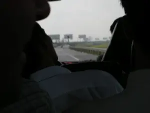 Bức ảnh được chụp từ bên trong một chiếc xe, có thể là ô tô hoặc xe buýt, đang di chuyển trên đường. Ở phía trước là bóng của những hành khách, trong đó có một người mặc áo sơ mi trắng. Nhìn qua kính chắn gió, có thể thấy một con đường quốc lộ hoặc đường cao tốc trải nhựa. Con đường có lan can bảo vệ ở bên cạnh. Xa xa trên đường có một chiếc xe máy đang chạy. Dọc theo con đường là một dải đất trồng cây xanh. Ở phía chân trời, có nhiều tấm biển quảng cáo lớn được dựng lên, nhưng do khoảng cách và độ mờ của ảnh nên không thể đọc được nội dung trên đó. Khung cảnh cho thấy một chuyến đi trên đường bộ, với thời tiết có vẻ u ám hoặc nhiều mây.。（日本語訳）。写真は、道路を走行中の車内、おそらく乗用車かバスの中から撮影されたものです。前方には乗客の影があり、その中には白いシャツを着た人が一人います。フロントガラス越しに、舗装された国道または高速道路が見えます。道路の脇にはガードレールがあります。道路の遠くには、一台のバイクが走っています。道路に沿って、緑の木々が植えられた土地が続いています。この光景は、曇りまたは雲の多い天気の中でのドライブ旅行を示しています。