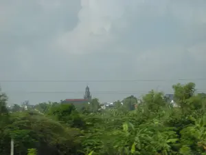 Bức ảnh chụp một khung cảnh vùng nông thôn, có thể được nhìn từ một phương tiện đang di chuyển. Tiền cảnh là một thảm thực vật xanh tươi và rậm rạp với nhiều loại cây cối. Ở phía xa, nổi bật giữa những lùm cây và các ngôi nhà là một công trình kiến trúc lớn, dường như là một nhà thờ Công giáo với tháp chuông cao và mái ngói đỏ. Bầu trời u ám, đầy mây. Một vài đường dây điện chạy ngang qua phần trên của bức ảnh. Không có hoạt động cụ thể nào của con người được nhìn thấy, chỉ có một khung cảnh tĩnh lặng của một ngôi làng hoặc vùng ngoại ô.。（日本語訳）。この写真は田舎の風景を撮影したもので、移動中の乗り物から見たものと思われます。前景には、様々な種類の木々が生い茂る、青々とした草木が広がっています。遠くには、木々や家々の間に、高い鐘楼と赤い瓦屋根を持つカトリック教会と思われる大きな建造物が際立っています。空はどんよりとしており、雲に覆われています。写真の上部を数本の電線が横切っています。特定の人間の活動は見られず、村または郊外の静かな風景だけが広がっています。