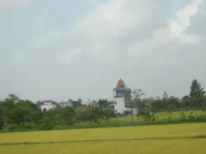 Bức ảnh ghi lại một phong cảnh nông thôn, có thể là ở khu vực Ninh Bình. Tiền cảnh là một cánh đồng lúa rộng lớn đang chín vàng, chuẩn bị cho mùa thu hoạch. Phía sau cánh đồng là một khu dân cư hoặc một ngôi làng với nhiều cây cối xanh tươi. Trong làng, có nhiều ngôi nhà với kiến trúc khác nhau. Nổi bật nhất là một tòa nhà cao tầng màu trắng và xanh, có một tháp với mái vòm màu đỏ. Ở phía xa hơn, có thể thấy một tòa nhà khác nhỏ hơn, màu hồng, cũng có kiến trúc mái vòm tương tự. Bầu trời trong ảnh có nhiều mây và khá u ám. Không có hoạt động nào của con người được nhìn thấy rõ. Không có chữ viết hay biển hiệu nào trong ảnh.。（日本語訳）。写真は、おそらくニンビン地方の田園風景を記録したものです。前景には、収穫の時期を迎え、黄金色に熟した広大な水田が広がっています。水田の後ろには、緑豊かな木々が生い茂る住宅地または村があります。村の中には、様々な建築様式の家が数多くあります。最も目立つのは、白と青の高層ビルで、赤いドーム屋根の塔があります。さらに遠くには、より小さなピンク色の建物が見え、それも同様のドーム屋根の建築様式です。写真の空は雲が多く、かなり曇っています。人間の活動ははっきりと見えません。写真には文字や看板はありません。