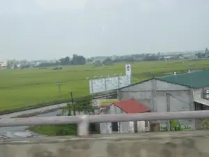 Bức ảnh chụp một khung cảnh nông thôn ở Ninh Bình, nhìn từ trên cao, có thể là từ một cây cầu hoặc đường cao tốc. Phía trước là một phần lan can cầu bị mờ. Trọng tâm của bức ảnh là một cánh đồng lúa xanh rộng lớn trải dài đến tận chân trời. Chạy dọc theo cánh đồng là một đường ray xe lửa và một con đường nhựa. Có một vài ngôi nhà nằm ven đường, bao gồm một ngôi nhà lớn có tường bê tông, mái tôn màu xanh lá cây và một ngôi nhà nhỏ hơn có mái ngói màu đỏ. Có một số biển quảng cáo, trong đó có một biển quảng cáo lớn với khung màu trắng chưa có nội dung, và một biển quảng cáo nhỏ hơn màu vàng và đỏ có logo của hãng Shell. Phía trên biển quảng cáo lớn còn có một biển hiệu khác với logo hình tròn màu đỏ. Các cột điện cũng xuất hiện rải rác trong khung cảnh. Bầu trời u ám và có nhiều mây.。（日本語訳）。ニンビンの田園風景を、おそらく橋か高速道路の上から撮影した写真です。手前には、ぼやけた橋の欄干の一部が見えます。写真の中心は、地平線まで広がる広大な緑の田んぼです。田んぼに沿って、線路とアスファルトの道路が走っています。道端にはいくつかの家があり、その中にはコンクリートの壁と緑色のトタン屋根を持つ大きな家と、赤い瓦屋根を持つ小さな家が含まれています。いくつかの広告看板があり、その中には内容がまだ書かれていない白い枠の大きな広告看板と、シェル社のロゴが入った黄色と赤の小さな広告看板があります。大きな広告看板の上には、赤い円形のロゴが入った別の看板もあります。電柱も風景の中に点在しています。空はどんよりと曇っています。