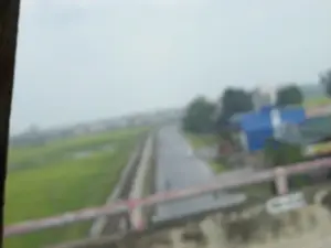 Bức ảnh rất mờ, được chụp từ trên cao, có thể là từ một cây cầu, nhìn xuống một khung cảnh nông thôn ở Việt Nam. Một con đường hoặc đường ray xe lửa chạy thẳng vào phía xa. Bên trái con đường là một cánh đồng lúa xanh mướt. Bên phải là một vài ngôi nhà, trong đó có một ngôi nhà có mái màu xanh lam, xen lẫn với cây cối. Ở tiền cảnh, có thể thấy lan can của cây cầu. Bầu trời u ám, cho thấy thời tiết có mây hoặc sắp mưa. Do chất lượng ảnh kém, không thể đọc được bất kỳ văn bản nào hoặc xác định các hoạt động cụ thể của con người.。（日本語訳）。写真は非常にぼやけており、上から、おそらく橋から撮影されたもので、ベトナムの田舎の風景を見下ろしています。一本の道または線路が遠方へとまっすぐに伸びています。道の左側には、青々とした水田が広がっています。右側には数軒の家があり、その中には青い屋根の家も一軒あり、木々に混じっています。前景には、橋の欄干が見えます。空はどんよりとしており、曇りまたは雨が降りそうな天気であることを示しています。写真の品質が悪いため、文字を読み取ったり、特定の人の活動を特定したりすることはできません。