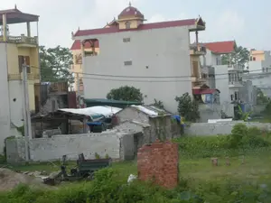 Bức ảnh mô tả một khung cảnh tại một khu dân cư, có thể là ở vùng ngoại ô hoặc nông thôn tại Ninh Bình. Phía sau là những ngôi nhà ống nhiều tầng được xây dựng san sát nhau với kiến trúc đa dạng, một số có mái ngói đỏ và các chi tiết trang trí trên đỉnh. Phía trước là một khu đất trống đang trong quá trình xây dựng. Một bức tường thấp làm từ gạch block xi măng được xây dựng dở dang. Gần đó có một chiếc xe công nông ba bánh (xe ba gác) dùng để chở vật liệu và một đống cát. Cỏ dại và cây cối mọc um tùm trên khu đất. Nhiều đường dây điện giăng ngang qua các tòa nhà. Không có hoạt động nào của con người được ghi lại, nhưng các vật thể cho thấy đây là một công trường xây dựng.。（日本語訳）。この写真は、おそらくニンビン省の郊外または田舎にある住宅地の一場面を描写しています。背景には、多様な建築様式で密集して建てられた多層階のチューブハウス（縦長の家）があり、中には赤い瓦屋根や頂部に装飾的な細部を持つものもあります。手前には、建設中の空き地があります。セメントブロックで作られた低い壁が、建設途中の状態です。その近くには、資材を運ぶための三輪の農業用車両（セ・バー・ガック）と砂の山があります。敷地には雑草や木々が生い茂っています。多くの電線が建物の間を横切っています。人間の活動は記録されていませんが、これらの物体はここが建設現場であることを示しています。