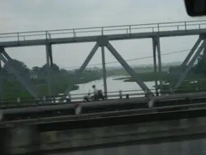 Bức ảnh được chụp từ một phương tiện đang di chuyển, ghi lại cảnh một cây cầu giàn bằng thép. Một người đang lái xe máy đi trên cây cầu. Nhìn qua kết cấu của cây cầu, có thể thấy một con sông chảy qua khu vực nông thôn với những cánh đồng xanh tươi, có thể là đồng lúa. Ở phía xa có cây cối và một vài ngôi nhà. Bầu trời u ám và có nhiều mây.。（日本語訳）。写真は移動中の乗り物から撮影されたもので、鋼鉄製のトラス橋の光景を捉えている。一人の人がバイクを運転して橋を渡っている。遠くには木々や数軒の家がある。空はどんよりとしており、雲が多い。