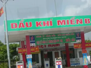 Bức ảnh chụp một trạm xăng dầu của PV OIL tại Việt Nam. Phía trên mái che là tấm biển lớn màu xanh lá cây với dòng chữ "DẦU KHÍ MIỀN BẮC" màu đỏ. Bên dưới là các biển hiệu khác cung cấp thông tin chi tiết: "CÔNG TY XĂNG DẦU - DẦU KHÍ MIỀN BẮC", "DOANH NGHIỆP NHÀ NƯỚC", và "ĐẠI LÝ BÁN LẺ XĂNG DẦU PV OIL - MIỀN BẮC". Logo của "PETROVIETNAM" và "PV OIL" cũng được hiển thị. Các trụ bơm xăng được đặt dưới mái che, và trên các cột trụ màu đỏ có các biển báo nhỏ cho biết loại nhiên liệu như "DIESEL", "XĂNG 92" và cảnh báo an toàn "CẤM LỬA". Ở phía bên trái, lá cờ Việt Nam đang bay trên một cột cờ. Phía sau có một băng rôn với dòng chữ "KHAI TRƯƠNG - KHUYẾN MẠI", cho thấy trạm xăng có thể mới mở hoặc đang có chương trình khuyến mãi. Khung cảnh xung quanh có cây cối và bầu trời có mây.。（日本語訳）。ベトナムにあるPV OILのガソリンスタンドを写した写真です。屋根の上には、赤色で「DẦU KHÍ MIỀN BẮC」と書かれた大きな緑色の看板があります。その下には、「CÔNG TY XĂNG DẦU - DẦU KHÍ MIỀN BẮC」、「DOANH NGHIỆP NHÀ NƯỚC」、そして「ĐẠI LÝ BÁN LẺ XĂNG DẦU PV OIL - MIỀN BẮC」といった詳細情報を提供する他の看板があります。「PETROVIETNAM」と「PV OIL」のロゴも表示されています。給油機は屋根の下に設置されており、赤い柱には「DIESEL」、「XĂNG 92」といった燃料の種類や、「CẤM LỬA」という安全警告を示す小さな標識があります。左側では、ベトナムの国旗が旗竿に掲げられてはためいています。後ろには「KHAI TRƯƠNG - KHUYẾN MẠI」と書かれた横断幕があり、このガソリンスタンドが新規開店したか、またはプロモーション中であることを示しています。周囲の風景には木々があり、空は曇っています。