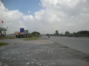 Bức ảnh chụp một khung cảnh ven đường quốc lộ ở Việt Nam. Bầu trời nhiều mây. Bên phải là một con đường nhựa rộng lớn với dải phân cách bằng kim loại, có các phương tiện giao thông như xe tải đang di chuyển ở phía xa và một người đang đi xe đạp ở lề đường. Bên trái là một khu vực đất đá và một trạm xăng. Có một số biển hiệu và cờ có thể nhận dạng được: một biển hiệu màu xanh lá cây của trạm xăng ghi 'DẦU KHÍ MIỀN BẮC'; một biển báo màu xanh dương với logo của PetroVietnam, ghi chữ 'PETROVIETNAM' và 'TCT DẦU VN'; một biển hiệu khác màu xanh lá cây ghi 'NHÀ NGHỈ 71' và 'VIỆT NAM'. Hai lá cờ đỏ sao vàng của Việt Nam đang được treo ở trạm xăng. Xung quanh có cây cối, nhà cửa và các cột điện.。（日本語訳）。写真はベトナムの国道沿いの風景を撮影したものです。空は曇っています。右側には金属製の中央分離帯がある広いアスファルトの道路があり、遠くにはトラックなどの車両が走行し、路肩では一人が自転車に乗っています。左側には土と石のエリアとガソリンスタンドがあります。いくつか識別できる看板と旗があります。ガソリンスタンドの緑色の看板には「DẦU KHÍ MIỀN BẮC」と書かれています。ペトロベトナムのロゴが入った青い看板には、「PETROVIETNAM」と「TCT DẦU VN」の文字が書かれています。別の緑色の看板には「NHÀ NGHỈ 71」と「VIỆT NAM」と書かれています。2つのベトナムの金星紅旗がガソリンスタンドに掲げられています。周囲には木々、家々、電柱があります。