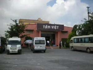 Bối cảnh là một bãi đậu xe phía trước một tòa nhà một tầng, sơn màu hồng, có thể là một trạm dừng chân hoặc một cơ sở kinh doanh. Trên mái hiên của tòa nhà có một biển hiệu lớn màu nâu với dòng chữ trắng 'TÂM VIỆT'. Trong bãi đậu xe có một số phương tiện đang đỗ, bao gồm một chiếc xe van Ford Transit màu trắng, một chiếc xe van màu bạc và một chiếc xe buýt nhỏ màu trắng và be. Trên đuôi chiếc xe van màu bạc có ghi 'KIM TOURS' và địa chỉ trang web 'www.kimtours.com.vn'. Giữa hai chiếc xe van có một chiếc ô lớn che nắng có logo của thương hiệu nước khoáng 'La Vie'. Một vài người đang di chuyển gần lối vào tòa nhà, cho thấy đây có thể là một điểm dừng nghỉ cho khách du lịch. Cảnh vật xung quanh có cây cối và một cột điện. Bầu trời có nhiều mây.。（日本語訳）。背景は、ピンク色に塗られた1階建ての建物の前にある駐車場で、休憩所か商業施設である可能性があります。建物の庇の上には、「TÂM VIỆT」と白い文字で書かれた茶色い大きな看板があります。駐車場には、白いフォード・トランジットのバン1台、銀色のバン1台、白とベージュのミニバス1台を含む、何台かの車両が駐車しています。銀色のバンの後部には、「KIM TOURS」とウェブサイトのアドレス「www.kimtours.com.vn」が書かれています。2台のバンの間には、ミネラルウォーターブランド「La Vie」のロゴが入った大きな日よけの傘があります。何人かの人々が建物の入り口近くを移動しており、ここが旅行客のための休憩地点である可能性を示しています。周囲の景色には木々と電柱が1本あります。空は雲が多いです。