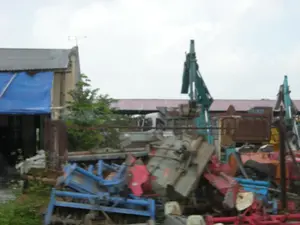 Bức ảnh chụp một khu vực ngoài trời, có vẻ là một bãi tập kết máy móc cũ hoặc một xưởng cơ khí. Ở phía trước là một đống lớn các thiết bị và phụ tùng máy móc nông nghiệp, chẳng hạn như máy cày, máy xới, với các màu sắc nổi bật như xanh lam và đỏ. Ở phía sau, có thể thấy các loại xe cơ giới hạng nặng, bao gồm ít nhất hai chiếc máy xúc màu xanh lục và một chiếc xe tải ben màu nâu đã gỉ sét. Một phần của một chiếc xe tải khác màu trắng cũng xuất hiện. Bên trái có một công trình xây dựng với mái lợp tôn, một phần được che bằng tấm bạt màu xanh. Trên mái nhà có một chiếc ăng-ten TV. Không có người trong ảnh và không có hoạt động nào đang diễn ra. Không có văn bản hay biển hiệu nào có thể đọc được.。（日本語訳）。写真は屋外のある場所を写しており、古い機械の集積場か、あるいは機械工場のように見えます。手前には、耕運機や鋤（すき）などの農業機械の設備や部品が山積みになっており、青や赤といった目立つ色をしています。後方には、少なくとも2台の緑色のショベルカーと、錆びた茶色のダンプトラックを含む、数種類の重機が見えます。白い別のトラックの一部も見えています。左側にはトタン屋根の建物があり、その一部は青い防水シートで覆われています。屋根の上にはテレビアンテナがあります。写真には人はおらず、何の活動も行われていません。読み取れる文字や看板はありません。