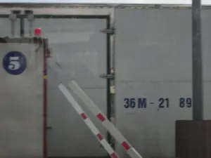 Bức ảnh chụp một phần của một chiếc xe tải lớn hoặc một container hàng hóa. Bề mặt xe có màu xám bạc, làm bằng kim loại. Ở bên trái, có một biển báo hình tròn màu xanh lam, bên trong có số "5" màu trắng. Phía trên biển báo là một chiếc đèn tín hiệu nhỏ màu đỏ. Ở phía trước, có hai thanh chắn barie, một thanh màu trắng và một thanh có sọc đỏ-trắng. Sự hiện diện của các thanh chắn này cho thấy đây có thể là một trạm kiểm soát, bến phà, hoặc khu vực bốc dỡ hàng hóa. Không có hoạt động nào của con người được nhìn thấy. Hình ảnh có vẻ được chụp từ bên trong một phương tiện khác nhìn ra ngoài qua cửa sổ.。（日本語訳）。写真は大型トラックまたは貨物コンテナの一部を撮影したものです。車両の表面はシルバーグレーで、金属製です。左側には、青い円形の標識があり、その中に白い数字の「5」があります。標識の上には、小さな赤い信号灯があります。前方には、白い遮断棒と赤と白の縞模様の遮断棒の2本の遮断機があります。これらの遮断機の存在は、ここが検問所、フェリー乗り場、または貨物の積み下ろしエリアである可能性を示唆しています。人間の活動は見られません。画像は、別の乗り物の内部から窓越しに撮影されたようです。