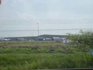 Bức ảnh chụp một khung cảnh ngoại ô, có khả năng là một công trường xây dựng lớn. Ở tiền cảnh là một khu đất trống mọc đầy cỏ dại và cây bụi, với hiệu ứng mờ cho thấy ảnh được chụp từ một phương tiện đang di chuyển. Ở phía xa hơn là một khu lán trại tạm bợ, có thể là nơi ở của công nhân, với nhiều ngôi nhà nhỏ được dựng lên sơ sài bằng các vật liệu đơn giản và được lợp bằng tấm bạt màu xanh và trắng. Phía sau khu lán trại là một bức tường bê tông cao và dài. Bầu trời có mây. Nhiều đường dây điện và một cột đèn đường bắc ngang qua khung cảnh. Về phía bên phải, có một tấm biển quảng cáo lớn màu trắng của một dự án. Tuy nhiên, các chữ viết trên đó bị mờ và không thể đọc rõ hoàn toàn, chỉ có thể nhận ra một vài ký tự như 'S...', 'TMA...', 'QUAT...', 'SQUARE'.。（日本語訳）。写真は郊外の風景を写しており、大きな建設現場である可能性があります。前景には、雑草や低木が生い茂る空き地があり、ぼかし効果から、移動中の乗り物から撮影されたことがわかります。さらに遠くには、労働者の居住地と思われる仮設のバラック地区があり、多くの小さな家が簡素な資材で粗末に建てられ、青と白の防水シートで屋根が覆われています。バラック地区の後ろには、高くて長いコンクリートの壁があります。空は曇っています。多くの電線と一本の街灯柱が風景を横切っています。右側には、あるプロジェクトの大きな白い広告看板があります。しかし、その上の文字はぼやけていて完全にはっきりと読むことはできず、「S...」、「TMA...」、「QUAT...」、「SQUARE」のような幾つかの文字しか認識できません。