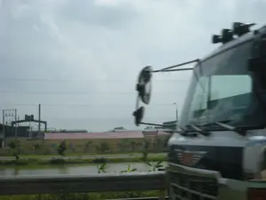 Bức ảnh được chụp từ một phương tiện đang di chuyển, nhìn ra khung cảnh bên đường. Ở phía bên phải, một phần đầu của một chiếc xe tải lớn đang chạy song song, có thể nhận ra logo của hãng xe Hino trên lưới tản nhiệt. Ở phía trước là lan can bảo vệ bằng kim loại của con đường. Phía sau lan can là một con kênh hoặc ao nhỏ. Bên kia bờ kênh là một bức tường dài màu vàng, phía sau là một tòa nhà dài có mái màu đỏ. Ở phía xa hơn là các công trình của một nhà máy hoặc khu công nghiệp với các cột và đường dây điện. Bầu trời u ám và đầy mây. Không có văn bản nào có thể đọc được, mặc dù có thể có chữ viết trên bức tường màu vàng nhưng đã bị mờ.。（日本語訳）。写真は移動中の乗り物から撮影されたもので、道端の風景が見えます。右側には、並走している大型トラックの先端部分があり、ラジエーターグリルには日野自動車のロゴが確認できます。前方には、道路の金属製のガードレールがあります。ガードレールの後ろには、水路または小さな池があります。水路の向こう岸には長い黄色の壁があり、その背後には赤い屋根の長い建物があります。さらに遠くには、電柱や電線のある工場または工業団地の建造物があります。空はどんよりとしており、雲に覆われています。読み取れる文字はありませんが、黄色の壁には文字が書かれているものの、ぼやけている可能性があります。