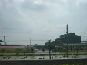 Bức ảnh chụp một khu công nghiệp hoặc nhà máy. Ở phía bên phải, có một tòa nhà lớn màu xanh đậm với một biển hiệu. Trên biển hiệu có các ký tự Trung Quốc "大量收购废铁" (nghĩa là: Thu mua phế liệu sắt số lượng lớn). Bên dưới có dòng chữ tiếng Việt nhưng khó đọc, có thể nhận ra các từ như "CÔNG TY TNHH..." và "THU MUA". Phía trước tòa nhà này có một lá cờ Việt Nam đang tung bay. Xung quanh là các tòa nhà nhà xưởng khác, một số có mái đỏ, và một ống khói công nghiệp ở phía xa bên trái. Toàn bộ khu vực được bao quanh bởi một bức tường màu vàng. Ở tiền cảnh, có một con kênh hoặc ao nước với một vài cây chuối nhỏ mọc ven bờ. Nhiều cột điện và đường dây điện chạy ngang qua khung cảnh. Phần dưới cùng của hình ảnh có một lan can, cho thấy ảnh được chụp từ một con đường hoặc cây cầu. Bầu trời có nhiều mây và không có hoạt động rõ ràng của con người trong ảnh.。（日本語訳）。写真は工業団地または工場を写したものです。右側には、看板のある大きな濃い青色の建物があります。看板には中国語の文字「大量收购废铁」（意味：鉄くずを大量に買い取ります）があります。その下にはベトナム語の文字がありますが、読みにくく、「CÔNG TY TNHH...」や「THU MUA」といった単語が認識できます。この建物の前には、ベトナムの国旗がはためいています。周囲には他の工場の建物があり、いくつかは赤い屋根で、左奥には工場の煙突があります。全域は黄色い壁で囲まれています。手前には、水路または池があり、岸辺には数本の小さなバナナの木が生えています。多くの電柱と電線が風景を横切っています。写真の最下部には欄干があり、写真が道路または橋から撮影されたことを示しています。空は雲が多く、写真には明確な人の活動は見られません。