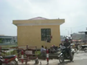 Bức ảnh mờ chụp một cảnh tại một điểm giao cắt đường sắt. Ở trung tâm là một chòi gác đường sắt sơn màu vàng. Phía trước có các thanh chắn đường sắt màu đỏ và trắng. Một người đội mũ bảo hiểm đang điều khiển xe máy ở phía trước bên phải, trong khi một số người khác đang ngồi gần đường ray, có vẻ như đang chờ tàu đi qua. Nhiều xe máy khác cũng đang dừng lại. Do ảnh bị mờ, không thể đọc được bất kỳ chữ viết nào trên các biển báo hay tòa nhà.。（日本語訳）。ぼやけた写真は、踏切の光景を写しています。中央には黄色く塗られた踏切の監視小屋があります。その手前には赤と白の遮断機があります。右手前ではヘルメットをかぶった人がバイクを運転しており、他の数人が線路の近くに座っており、電車が通過するのを待っているようです。他にも多くのバイクが停止しています。写真がぼやけているため、標識や建物に書かれている文字は一切読み取れません。