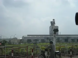 Trên bức tường của một tòa nhà lớn giống như nhà máy, có dòng chữ màu xanh lam: 'NHÀ MÁY THỨC ĂN CHĂN NUÔI DANMACH'. Bối cảnh là một khu vực công nghiệp hoặc ngoại ô vào một ngày trời nhiều mây. Ở phía trước, có một đường ray xe lửa với rào chắn ngang đường màu đỏ và trắng. Một người đang ngồi trên xe máy, đội mũ bảo hiểm, dường như đang dừng lại chờ tàu hỏa đi qua. Phía sau rào chắn là nhà máy lớn và một số tòa nhà phụ khác. Dọc theo con đường ở phía xa bên trái là một hàng cột đèn đường.。（日本語訳）。工場のような大きな建物の壁には、「デンマーク飼料工場」という青い文字があります。背景は、曇りの日の工業地帯または郊外です。手前には、赤と白の踏切遮断機がある線路があります。ヘルメットをかぶった人がバイクに乗っており、列車が通過するのを待って停止しているようです。遮断機の後ろには、大きな工場と他のいくつかの付属の建物があります。左奥の道沿いには、街灯の列があります。