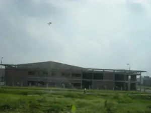 Bức ảnh chụp một công trình xây dựng lớn, hai tầng, đang trong giai đoạn thi công dở dang. Tòa nhà có kết cấu khung bê tông và tường gạch, nhưng chưa được lắp cửa sổ hay hoàn thiện. Phía trước tòa nhà là một khu đất rộng với cỏ xanh mọc um tùm, có thể là một cánh đồng. Xung quanh công trình có một vài cột đèn đường. Bầu trời u ám, nhiều mây. Hình ảnh có vẻ được chụp từ bên trong một phương tiện đang di chuyển, qua một tấm kính có vài vết bẩn. Không có văn bản hay chữ viết nào có thể đọc được trong ảnh.。（日本語訳）。写真は、建設途中の大きな2階建ての建設現場を写している。建物はコンクリートの骨組みとレンガの壁でできているが、まだ窓は取り付けられておらず、完成もしていない。建物の前には、青々とした草が生い茂る広い土地があり、野原かもしれない。建設現場の周りには、いくつかの街灯がある。空は曇っており、雲が多い。写真は、移動中の乗り物の中から、いくつかの汚れが付いたガラス越しに撮影されたようだ。写真の中には、読み取れる文字や文章はない。