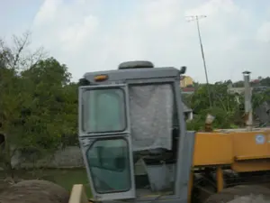 写真は、農村地帯の建設中と思われる場所で、重建設車両の一部を撮影したものです。それはブルドーザーか道路舗装機かもしれません。車両の運転室は灰色で、ガラスのドアが開いています。車両の後部は黄色です。背景には、たくさんの青々とした木々と、小さな運河か池のほとりにある土手が含まれています。さらに遠くには民家があり、その中には黄色く塗られた家と、曇り空に向かって高く伸びるテレビアンテナがあります。前景、車両の近くには、土か砂の大きな山があり、ここが建設現場である可能性を示唆しています。写真には読み取れる文字や記号はありません