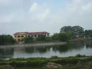 Bức ảnh chụp một khung cảnh ven một hồ nước hoặc một con sông. Nổi bật trong ảnh là một tòa nhà lớn hai tầng nằm bên bờ nước. Tòa nhà này có kiến trúc đặc trưng, có thể là một công trình công cộng như nhà văn hóa hoặc trụ sở cơ quan, với tường sơn màu vàng và mái lợp ngói đỏ. Phía trước tòa nhà có treo một lá cờ Việt Nam. Xung quanh tòa nhà và dọc theo bờ kè bê tông là rất nhiều cây xanh. Mặt nước phẳng lặng, in bóng tòa nhà và cây cối. Phía trước, gần với người chụp ảnh, là một bờ đất với cỏ mọc. Bầu trời có nhiều mây. Không có người hay hoạt động nào được ghi nhận trong ảnh, tạo nên một không gian yên tĩnh. Không có văn bản hay chữ viết nào có thể đọc được.。（日本語訳）。写真は、湖または川のほとりの風景を撮影したものです。写真の中で目立っているのは、水辺に建つ大きな2階建ての建物です。この建物は特徴的な建築様式で、文化会館や官公庁の庁舎のような公共施設である可能性があり、壁は黄色に塗られ、屋根は赤い瓦で葺かれています。建物の正面にはベトナムの国旗が掲げられています。建物の周りやコンクリートの護岸に沿って、たくさんの緑の木々があります。水面は穏やかで、建物や木々を映しています。手前、撮影者に近い側には、草の生えた土手があります。空には多くの雲があります。写真には人や活動は記録されておらず、静かな空間を作り出しています。読み取れるテキストや文字はありません。