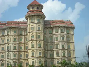 Bức ảnh chụp một tòa nhà lớn, nhiều tầng với kiến trúc phức tạp và trang trí công phu, có thể là một khách sạn. Tòa nhà có màu vàng kem, nổi bật với một tháp hình trụ ở góc và mái lợp ngói đỏ. Toàn bộ mặt tiền có rất nhiều cửa sổ hình vòm được sắp xếp đối xứng. Kiến trúc này là sự pha trộn giữa phong cách cổ điển châu Âu và các yếu tố Á Đông. Bối cảnh là bầu trời ban ngày trong xanh với vài đám mây trắng. Phía trước tòa nhà có ngọn cây cọ và một vài đường dây điện. Không có bất kỳ biển hiệu, chữ viết hay hoạt động nào của con người được ghi lại trong ảnh.。（日本語訳）。写真には、複雑な建築と精巧な装飾が施された、大きな多層階の建物が写っており、ホテルである可能性があります。建物はクリームイエロー色で、角にある円筒形の塔と赤い瓦屋根が際立っています。ファサード全体には、対称的に配置された多くのアーチ型の窓があります。この建築様式は、ヨーロッパの古典的なスタイルとアジアの要素の融合です。背景は、いくつかの白い雲が浮かぶ昼間の青空です。建物の前には、ヤシの木の先端と数本の電線があります。写真には、いかなる看板、文字、または人間の活動も記録されていません。