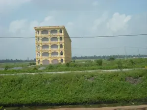 Bức ảnh chụp một cảnh quan ở vùng nông thôn, có thể là ở khu vực Ninh Bình. Nổi bật giữa khung cảnh là một tòa nhà lớn, cao khoảng 5 tầng, được sơn màu vàng. Tòa nhà này có thiết kế đối xứng với nhiều ban công và các ô cửa sổ, cửa ra vào hình vòm, nhưng có vẻ như nó chưa hoàn thiện hoặc bị bỏ hoang, đứng trơ trọi giữa một cánh đồng rộng lớn. Xung quanh tòa nhà là đất trống và cây cỏ dại mọc um tùm. Phía trước là một con đường đất. Ở tiền cảnh là thảm thực vật xanh tươi, có thể là một bờ mương hoặc một phần của cánh đồng. Ở phía sau và xa hơn là những cánh đồng khác, hàng cây, và các cột điện. Một đường dây điện chạy ngang qua phần trên của bức ảnh. Bầu trời trong xanh và có mây trắng. Không có người hay hoạt động nào được ghi nhận. Không có bất kỳ văn bản, biển báo nào trong ảnh.。（日本語訳）。写真は、おそらくニンビン地域の田園風景を撮影したものです。風景の中でひときわ目立っているのは、黄色に塗られた高さ約5階建ての大きな建物です。この建物は、多くのバルコニーとアーチ型の窓や出入り口を持つ左右対称のデザインですが、未完成または廃墟のようで、広大な野原の真ん中にぽつんと建っています。建物の周りは空き地で、雑草が生い茂っています。手前には未舗装の道があります。前景には青々とした植物があり、用水路の土手か畑の一部かもしれません。後方およびさらに遠くには、他の畑、並木、そして電柱があります。一本の電線が写真の上部を横切っています。空は青く澄んでおり、白い雲があります。人や活動の様子は記録されていません。写真にはいかなる文字や看板もありません。
