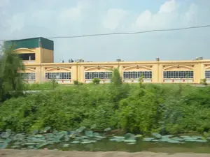 Bức ảnh chụp một tòa nhà lớn màu vàng, có thể là một nhà máy hoặc một công trình công cộng, nằm ở vùng nông thôn. Tòa nhà có một phần hai tầng với mái tôn màu xanh lá cây và phần còn lại là một dãy nhà một tầng với nhiều cửa sổ. Trên sân thượng của tòa nhà có một nhóm người, dường như là công nhân đang làm việc. Phía trước tòa nhà là một khu vực cây cối rậm rạp. Ở tiền cảnh là một ao hoặc kênh nước có trồng sen, với nhiều lá sen và một vài bông hoa sen màu hồng đang nở. Một đường dây điện chạy ngang qua bầu trời có mây. Không có văn bản hay biển hiệu nào có thể đọc được trong ảnh.。（日本語訳）。写真は、田舎にある、工場か公共施設と思われる大きな黄色い建物を写しています。建物は、緑色のトタン屋根を持つ2階建ての部分と、残りは多くの窓がある平屋建ての棟で構成されています。建物の屋上には、作業をしていると思われる労働者の一団がいます。建物の前には、木々が鬱蒼と茂るエリアがあります。前景には、蓮が植えられた池か水路があり、たくさんの蓮の葉と、いくつかのピンク色の蓮の花が咲いています。曇り空を電線が横切っています。写真の中には、読み取れる文字や看板はありません。