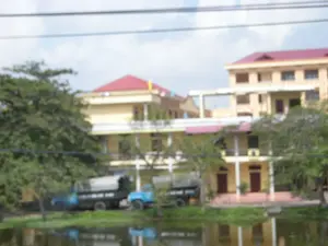 Bức ảnh bị mờ, chụp một khung cảnh ven một ao hoặc sông ở Ninh Bình. Ở phía sau, có một số tòa nhà nhiều tầng, sơn màu vàng với mái ngói màu đỏ. Phía trước các tòa nhà, có hai chiếc xe tải màu xanh lam đang đỗ bên cạnh nhiều cây xanh. Mặt nước ở phía trước phản chiếu hình ảnh của các tòa nhà và cây cối. Phía trên cùng của bức ảnh có các đường dây điện chạy ngang qua. Không có chữ viết nào có thể đọc được và không có hoạt động nào của con người được quan sát thấy trong ảnh.。（日本語訳）。写真はぼやけており、ニンビンの池か川のほとりの風景を写しています。後方には、いくつかの多層階の建物があり、黄色く塗られ、赤い瓦屋根です。建物の前には、2台の青いトラックが多くの緑の木の隣に停まっています。前方の水面は、建物や木々の姿を映しています。写真の最上部には電線が横切っています。読み取れる文字はなく、写真の中には人間の活動は見られません。